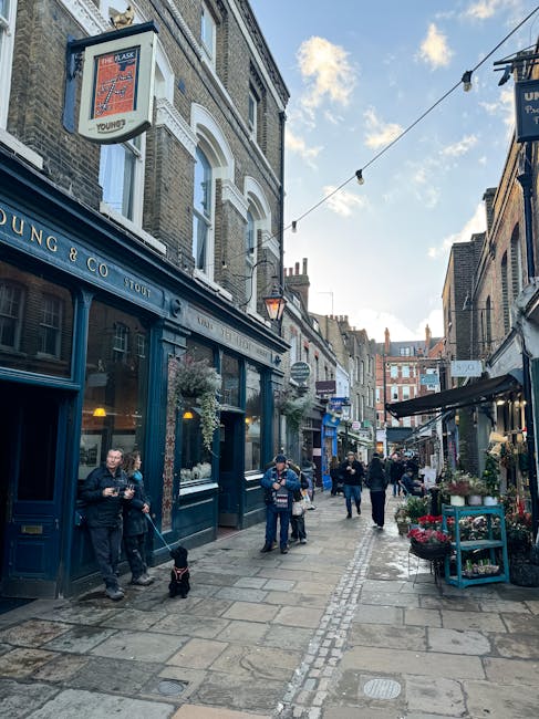 A narrow street scene in Hampstead, featuring a row of traditional brick and stone buildings with shops, pubs, and cafes, some with hanging flower baskets and outdoor displays. The pavement is lined with pedestrians, including a man and woman standing outside a blue pub, Hampstead & Co., with a dog on a leash. Several individuals are walking along the street, some shopping or socialising. Overhead, string lights are strung across the street, and the sky is partly cloudy with patches of blue. The environment suggests a lively area suitable for house removals or home relocation services, with visible commercial properties and pedestrian activity, providing context for moving and packing logistics operated by Hampstead Man and Van.