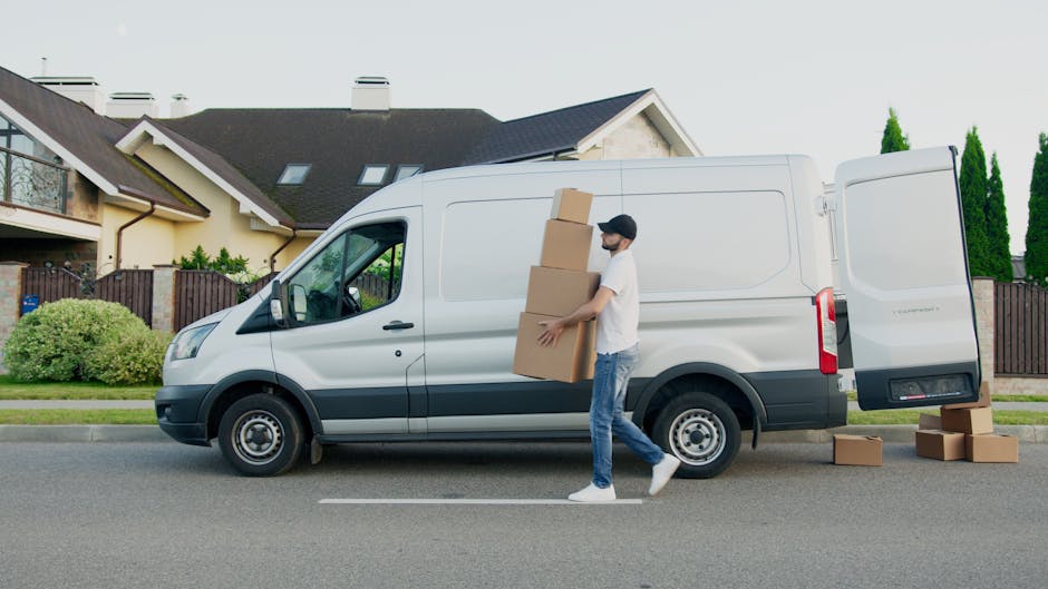 A man wearing a white t-shirt, blue jeans, and a black cap is next to a white panel van parked on a residential street, carrying three large cardboard boxes stacked vertically. Additional cardboard boxes are placed on the pavement beside the van, with some stacked near the open rear door. The van is adjacent to a suburban house with a pitched roof, dormer windows, and a wooden gate, and is surrounded by greenery including bushes and tall trees. The scene appears to be part of a home relocation or furniture transport process, with the individual preparing to load or unload items as part of packing and moving activities. Hampstead Man and Van is involved in the vehicle and removals services relevant to this house move, with the environment lit by natural daylight.