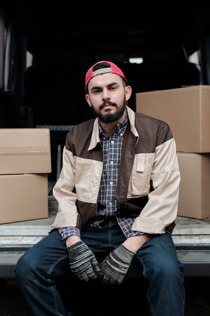 A man with a light beard and smiling face, wearing a blue knit beanie and a colour-blocked jacket in navy blue, red, and grey, is sitting on the edge of an open van with its rear doors wide open. Insi
