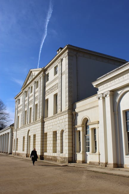A person walking across a gravel pathway outside a large classical-style building with ornate architectural details and decorative columns. The building's facade features tall windows, stucco walls, and intricate molding, with the upper section showing a slightly protruding tier with additional decorative elements. The scene is set on a clear day with a bright blue sky and a visible contrail. The environment suggests a historical or estate setting, possibly involved in house or estate relocation services. Hampstead Man and Van's removals team may be pictured preparing or assessing the site for furniture transport and packing during a home relocation process, with the building's spacious exterior indicating the capacity for large items and furniture loading and unloading nearby.