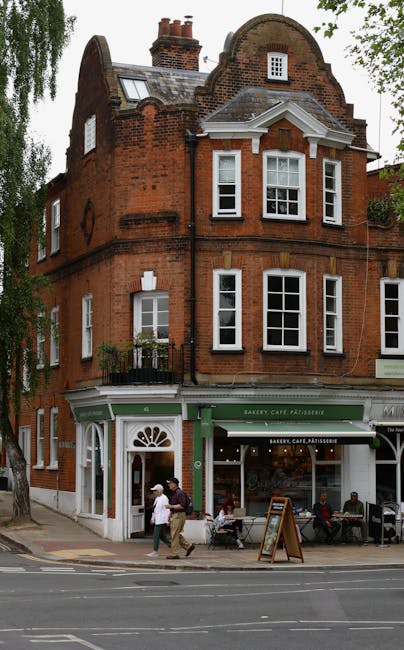 A multi-storey red brick building with a curved gable roof and bay windows on the upper floors, located on a street corner in Hampstead. The ground floor features a bakery, café, and patisserie with a green and white storefront, including a large front window, an entry door with a semi-circular window above, and an outdoor chalkboard menu sign. Several pedestrians are walking past the shop, with some seated outside on benches or chairs. The building's exterior showcases typical Victorian architecture, and there are trees visible around the structure. This scene is set during daytime with natural lighting, illustrating an active street environment suitable for home relocation or furniture transport considerations in the Hampstead area. Hampstead Man and Van is familiar with such urban settings for house removals and moving services.