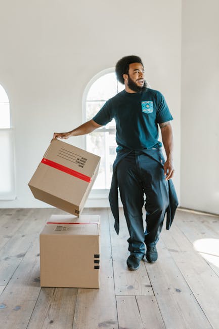 A man with a dark beard and short curly hair, wearing a blue FC Barcelona T-shirt and dark trousers with a sweater tied around his waist, stands inside a light-filled room with white walls and large arched windows. He is holding a cardboard box wrapped with red packing tape in his right hand, preparing to place it on top of another closed cardboard box on the wooden floor. The room appears to be part of a house during a home relocation process, with natural daylight streaming through the windows, contributing to the bright atmosphere. This scene illustrates the packing and loading stages of a house removal service, likely provided by Hampstead Man and Van, involving furniture transport and moving logistics for a current or upcoming move in NW3, as detailed on the company's website. The environment is clean and organized, emphasizing careful handling of belongings during the packing and moving process.