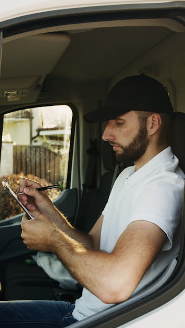 A man with a beard, wearing a black cap and a white polo shirt, seated inside a light-colored van, is engaged in a home relocation process. He is holding a smartphone in one hand and using a pen or stylus with the other to make notes or review information, likely related to the move. The van's interior is visible, showing the ceiling and part of the window, through which an outdoor area with a gate and some foliage can be seen. This scene captures a moment of planning or coordination during furniture transport or packing and moving activities, with the man possibly managing logistics for Hampstead Man and Van’s removal services. Behind him, the vehicle's driver’s seat and dashboard are partially visible, indicating that the van is in a stationary position, ready for further loading or unloading procedures typical in house removals or same-day relocation services.